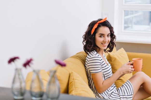 Charmante junge Frau im Kleid lächelnd auf der Couch in der modernen Wohnung. Orange, Weiß, goldene Farben, fröhliche Stimmung, Lächeln, Ausruhen, Entspannen