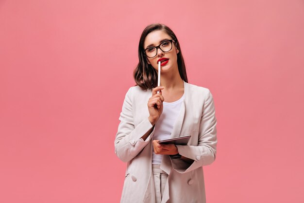 Charmante junge Frau mit dunklem Haar im klassischen Stil Anzug und Brille hält Computertablett auf lokalisiertem rosa Hintergrund.