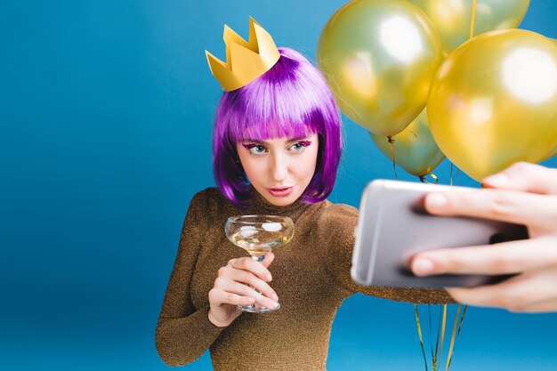 Charmante junge Frau mit lila Haarschnitt, Krone auf Kopf machen Selfie-Porträt. Goldene Luftballons, Champagner, Neujahrsparty, Luxuskleid, Lametta-Make-up.