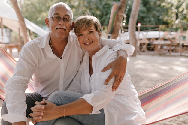 Charmante kurzhaarige Dame in Bluse und Jeans lächelnd, in die Kamera schauend und mit altem Mann in Brille und weißem Hemd am Strand umarmend.