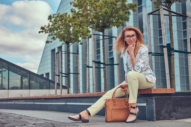 Charmante modische Frau in stilvoller Kleidung und Brille mit Handtasche, die auf einer Bank gegen einen Wolkenkratzer sitzt.