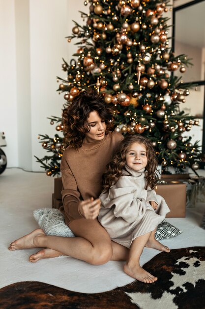 Charmante Mutter und Tochter mit lockiger Frisur haben Spaß, umarmen und küssen zu Hause in der Nähe des Weihnachtsbaumes in einem weißen Interieur