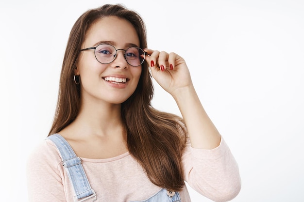 Charmante nerdige junge weibliche Brünette mit perfektem weißem Lächeln, die Hände auf der Brille hält und mit selbstbewusstem, glücklichem und selbstbewusstem Ausdruck lächelt, der über grauer Wand erfreut steht