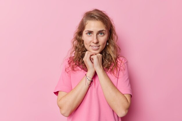 Charmante, schöne, blauäugige junge Frau mit langem, welligem Haar hält die Hände unter dem Kinn und schaut direkt in die Kamera, gekleidet in einem lässigen Basic-T-Shirt, das isoliert über rosafarbenem Hintergrund posiert, um Fotos zu machen