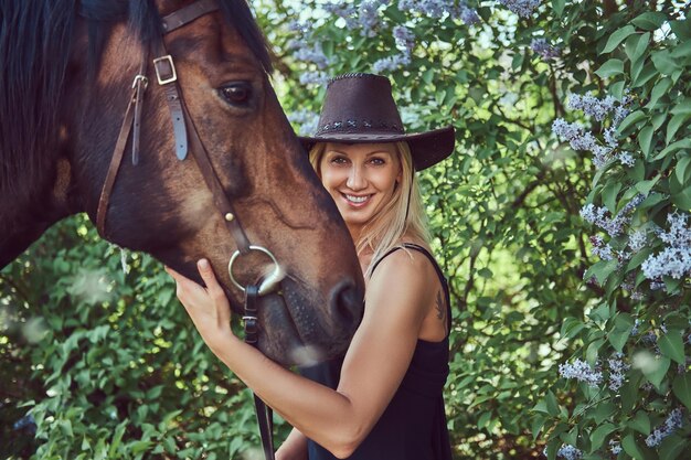Charmante schöne Blondine in schwarzer Kleidung und Hut, die ein Pferd im Blumengarten umarmt.