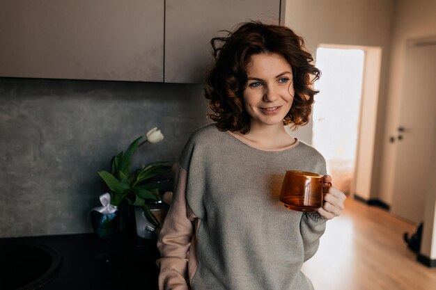 Charmante schöne Frau mit Locken im Hausanzug, die sich mit Tee in der Küche entspannt