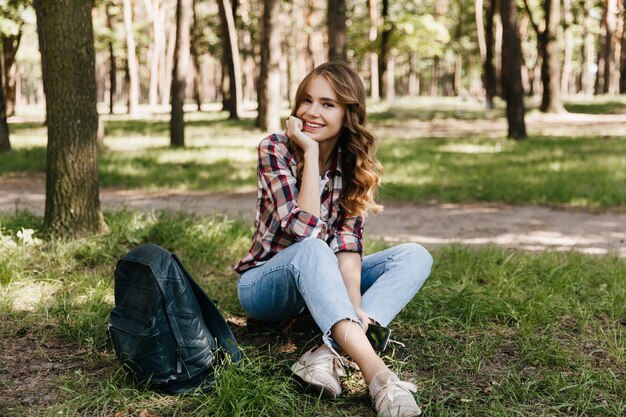 Charmante weibliche Touristin, die im Park mit Rucksack aufwirft. Außenaufnahme des erstaunlichen lockigen Mädchens, das auf dem Gras mit Lächeln sitzt.