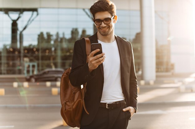 Charmanter brünetter Mann im schwarzen Anzug chattet am Telefon und lächelt Junger Mann mit Brille hält braunen Rucksack und posiert draußen