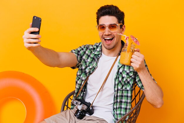 Charmanter Mann im karierten Hemd hält orange Cocktail und nimmt Selfie. Kerl in der Sonnenbrille wirft mit Retro-Kamera auf isoliertem Raum mit aufblasbarem Kreis auf.