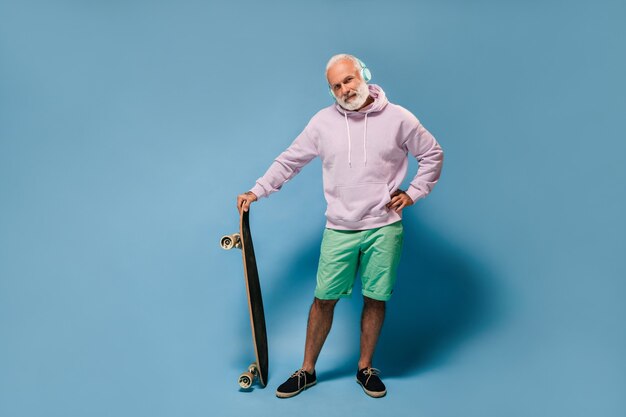 Charmanter Mann in Hoodie und grünen Shorts, der Musik hört und ein Skateboard hält