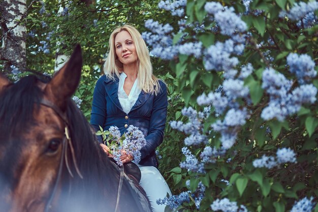 Charmanter schöner blonder Jockey, der auf einem braunen Pferd im Blumengarten reitet.