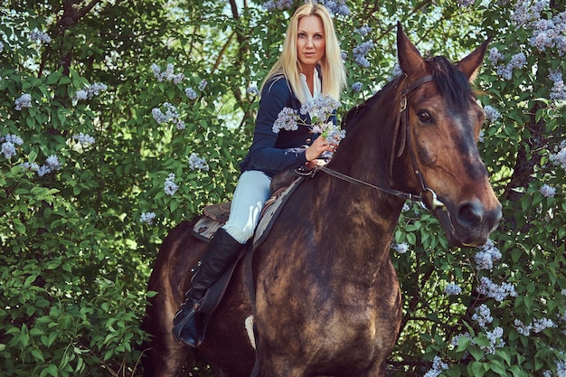 Charmanter schöner blonder Jockey, der auf einem braunen Pferd im Blumengarten reitet.