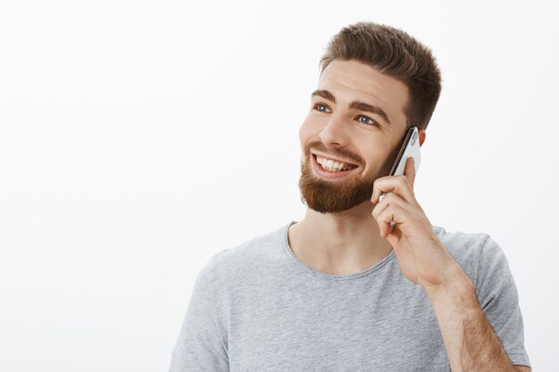 Charmanter sorgloser junger erfolgreicher Unternehmer mit blauen Augen und Bart, der Handy nahe Ohr hält, das links mit verträumtem freudigem Blick schaut und lässig über smarpthone über weiße Wand redend lächelt