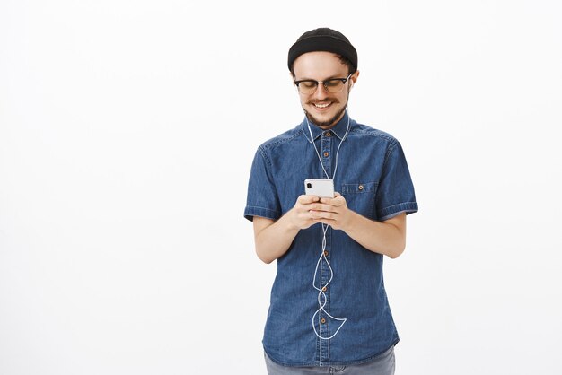 Charmanter und freudiger gutaussehender erwachsener Kerl in Mütze und Brille mit Bartnachrichten während der Reise in der U-Bahn, die Smartphone hält, das Musik in den Ohrhörern hört, zufrieden mit niedlicher romantischer Note