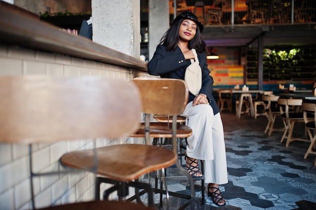 Charmantes afrikanisch-amerikanisches Frauenmodell mit schwarzem Jackenhut und Gürteltasche, das sich in der Freizeit im Café entspannt