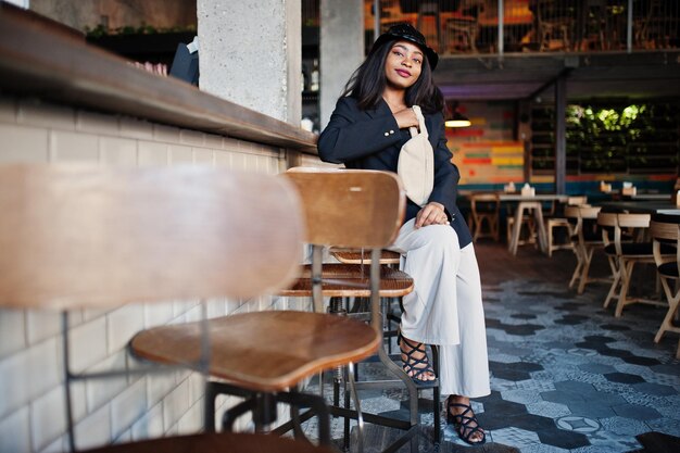 Charmantes afrikanisch-amerikanisches Frauenmodell mit schwarzem Jackenhut und Gürteltasche, das sich in der Freizeit im Café entspannt