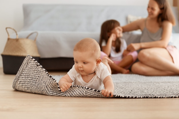 Charmantes Babymädchen, das die Welt studiert, Teppich berührt, während es im Wohnzimmer auf dem Boden liegt, entzückendes Kind, das weiße Kleidung trägt, Mutter und Schwester, die im Hintergrund posieren.