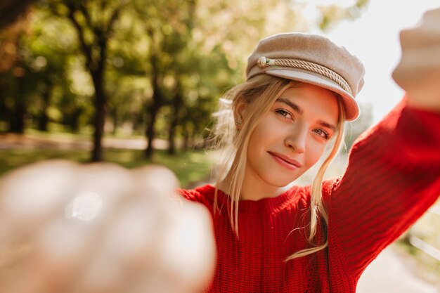 Charmantes blondes Mädchen im hellen trendigen Hut und im roten Pullover, der Selfie im Herbstpark macht.