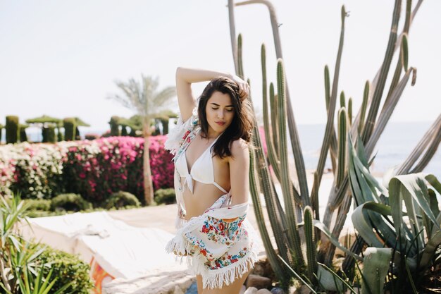 Charmantes dunkelhaariges Mädchen in weißen Kleidern mit ernstem Gesichtsausdruck, der neben Kaktus steht und Sommersonne genießt. Porträt der schlanken jungen brünetten Frau im Bikini, der auf exotischer Landschaft aufwirft