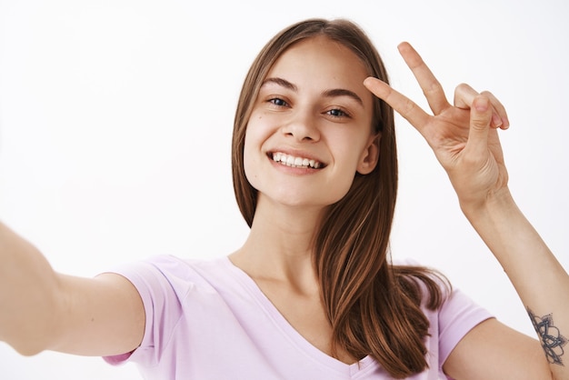 Charmantes, freundlich aussehendes, freudiges Mädchen mit braunem Haar, das breit lächelt und Frieden oder Siegeszeichen nahe Gesicht zeigt, während Selfie mit Smartphone über graue Wand nimmt