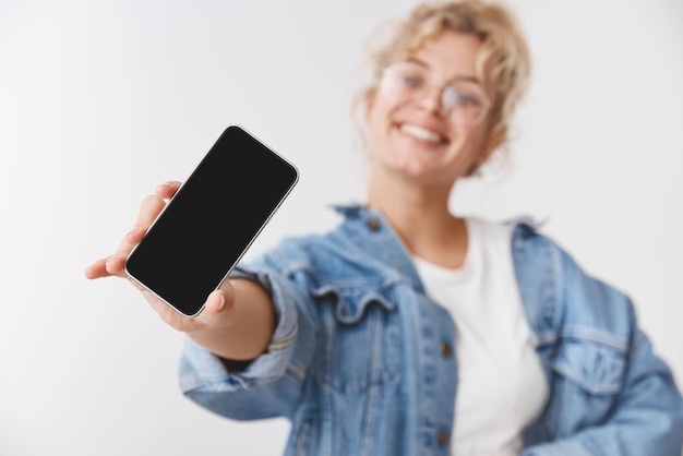 Charmantes, glückliches, gutaussehendes, energetisiertes, junges, blondes, kaukasisches Mädchen, das hinter dem Stretch-Handshow-Kamera-Smartphone-Display verschwommen ist und das Telefonanwendungs-Gadget auf weißem Hintergrund fördert
