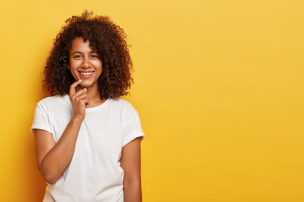 Charmantes, gut aussehendes junges Mädchen mit Afro-Haaren, lächelt sanft, hat natürliche Schönheit, ist in Hochstimmung, genießt eine tolle Zeit am Wochenende und trägt weiße Freizeitkleidung, die auf Gelb isoliert ist