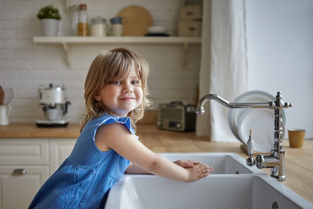 Charmantes kleines Mädchen im blauen Kleid, das Hände in der Küche wäscht. Nettes weibliches Kind, das Kamera betrachtet und lächelt, Mutter hilft, Abwasch macht, am Waschbecken steht. Kinder, Kindheit, Kochen und Hausarbeit