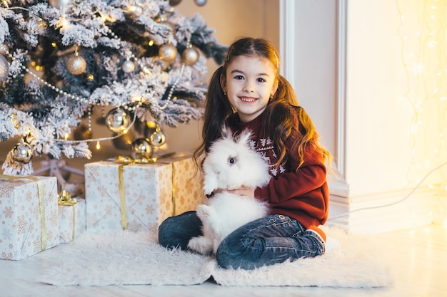 Charmantes kleines Mädchen sitzt mit einem Hasen vor einem glänzenden Weihnachtsbaum