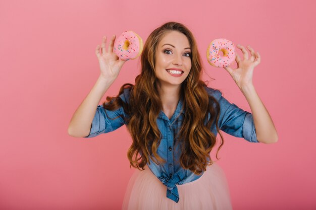 Charmantes langhaariges Mädchen im trendigen Outfit gebackene glasierte leckere Donuts zum Tee mit der Familie. Attraktive lockige junge Frau, die aufwirft, die köstliche Donuts vom Bäckereigeschäft lokalisiert auf rosa Hintergrund hält