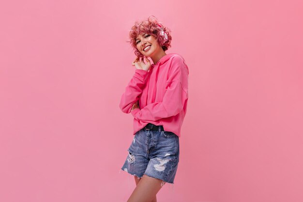 Charmantes lockiges Mädchen in Jeanshose und rosa Hoodie lächelt breit Porträt einer glücklichen Frau mit Kopfhörern, die Musik auf isoliertem Hintergrund hört