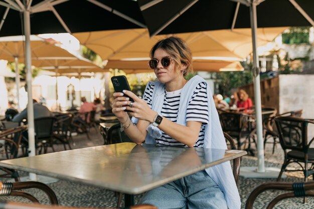 Charmantes Mädchen in Jeans und gestreiftem T-Shirt mit Sonnenbrille und Smartphone in den Händen sitzt im Café im Freien und wartet darauf, dass der Kellner eine Bestellung aufgibt Hübsche junge Frau kam zum Mittagessen