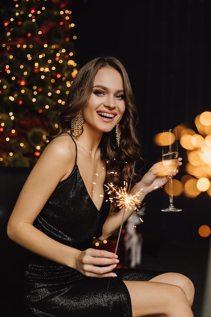 Charmantes Mädchen lächelt und hält Wunderkerze und ein Glas mit Champagner auf einer Neujahrsparty