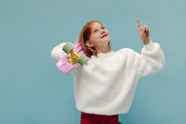 Charmantes Mädchen mit fuchsiger Frisur in weißem, trendigem Pullover und roter Hose, die auf Platz für Text zeigt und mit Skateboard auf blauem Hintergrund posiert