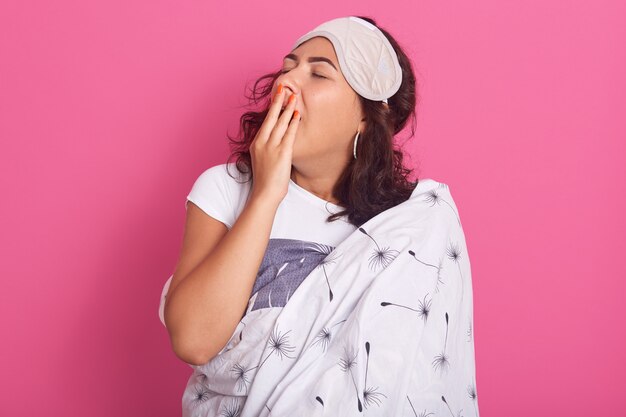 Charmantes Mädchen mit Schlafmaske und Decke umwickelt, bedeckt ihren offenen Mund mit der Hand beim Gähnen, Dame sieht schläfrig aus