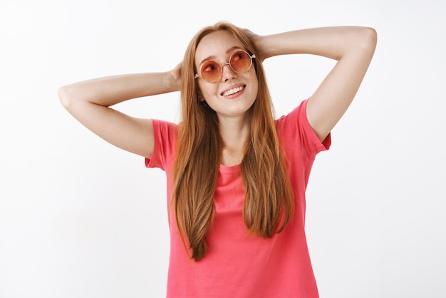 Charmantes sorgloses Hippie-Mädchen mit Ingwerhaar und Sommersprossen in der stilvollen rosa Sonnenbrille, die Hände hinter dem Kopf hält, der in der faulen Haltung steht und die obere rechte Ecke anstarrt