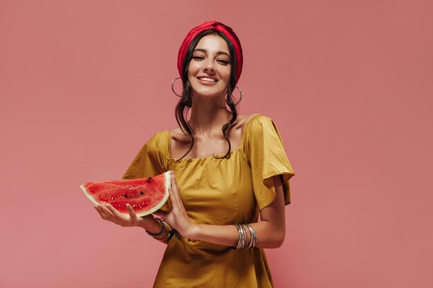 Charmantes stilvolles Mädchen mit welligem dunklem Haar in stilvollen Accessoires und gelbem Sommerkleid, das lächelt und Wassermelone auf rosa Hintergrund hält