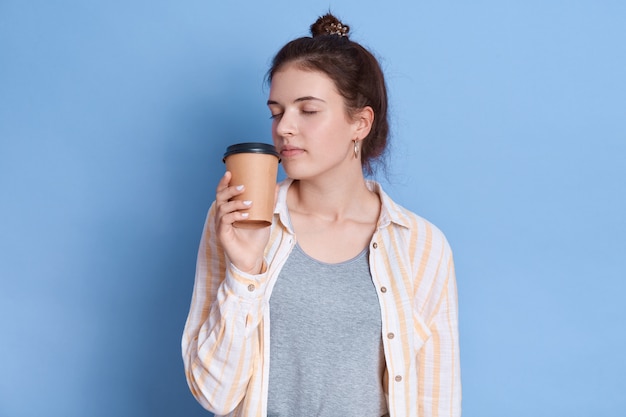 Charmantes süßes lässiges, dunkelhaariges, ziemlich faszinierendes Mädchen, das ihren Tee oder Kaffee in Pappbecher riecht und isolierte Freizeithemden trägt.