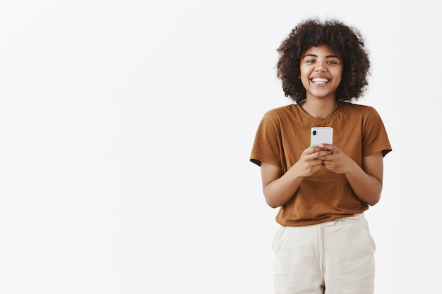 Charmantes und freundliches afroamerikanisches Teenager-Mädchen mit lockigem Haar im stilvollen Outfit, das Smartphone hält und breit lächelt