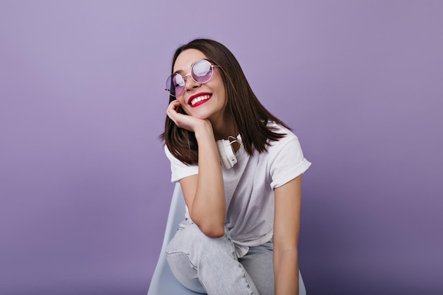 Charmantes weibliches Modell in Kopfhörern, das mit einem Lächeln über etwas nachdenkt Innenporträt eines glücklichen brünetten Mädchens isoliert auf violettem Hintergrund