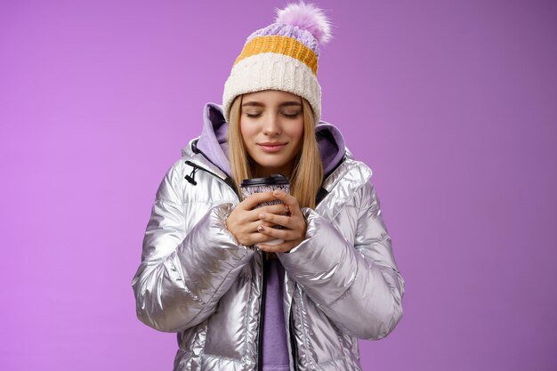 Charmantes zartes romantisches blondes Mädchen, das schönen Geruch warmer, leckerer Kaffee schnüffelt, der warme Hände zum Mitnehmen hält, schließen die Augen und sehen erfreut aus, wenn sie eine Winterjacke tragen, genießen Sie den Urlaub, lila Hintergrund.