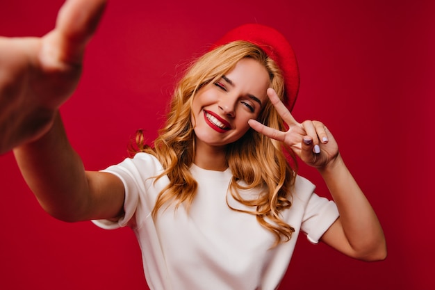 Cheerul gut aussehende Frau, die Selfie auf roter Wand macht. Innenaufnahme des stilvollen sorglosen französischen Mädchens lächelnd