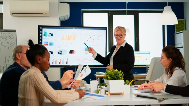 Chefanalyst, der eine Besprechungspräsentation für ein Team von Ökonomen hält. Manager, der ein digitales interaktives Whiteboard mit Wachstumsanalyse, Diagrammen, Statistiken, Daten und verschiedenen Personen zeigt, die in Broadroom arbeiten