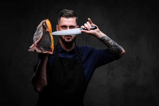 Chefkoch hält Messer und großes Stück exklusives Wurstwaren auf dunklem Hintergrund.