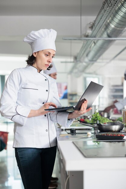 Chefköchin mit Laptop-Brainstorming garniert Ideen für Gourmetgerichte, während sie in der Restaurantküche steht. Professioneller Arbeiter in der Lebensmittelindustrie mit Computer, der nach gastronomischen Gerichten sucht.