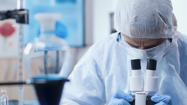 Chemiker-Forscher-Arzt, der eine PSA-Ausrüstung trägt, um eine Infektion mit Coronavirus zu verhindern, der eine Virusprobe mit einem Mikroskop analysiert, das im medizinischen Impfstoff im Chemielabor arbeitet. Medizinkonzept