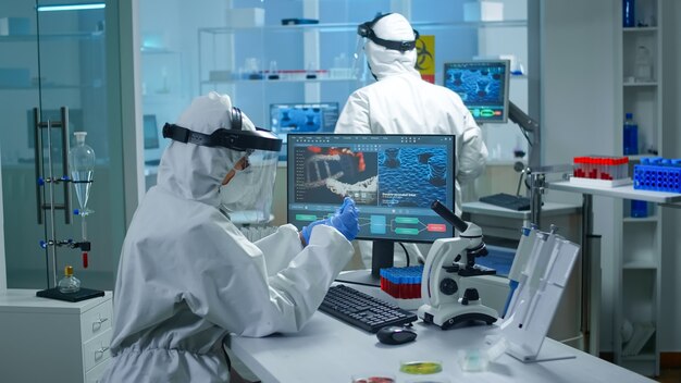 Chemiker im PPE-Anzug, der am PC arbeitet, während die Labortechnikerin ihre Blutproben bringt