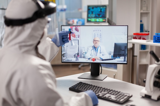 Chemiker im ppe-anzug hört professionellen arzt bei videoanrufen zu und diskutiert während eines virtuellen treffens im forschungslabor