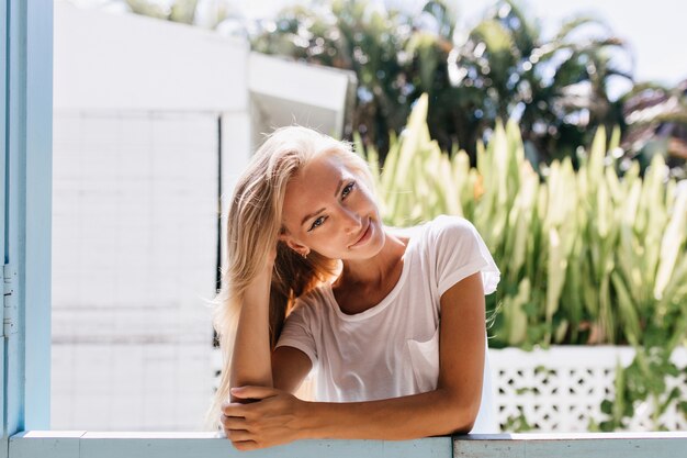 Chilling gebräunte Frau posiert mit schlauen Lächeln. Faszinierende junge Dame mit blondem glänzendem Haar, das Wochenende genießt.