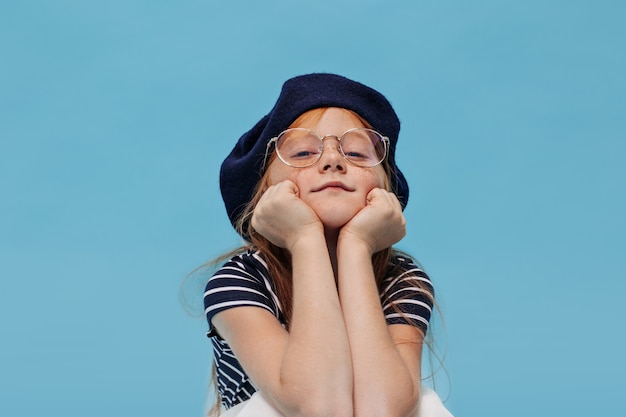 Cleveres kleines Mädchen mit Sommersprossen in stylischem Hut und klarer Brille posiert und schaut nach vorne auf blau isolierte Wand