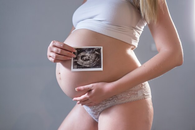 Close-up von Bauch der jungen schwangeren Modell zeigt Ultraschall-Bild von ihrem Baby. Zukünftige Mama auf ihrem zweiten Trimester mit Ultraschalluntersuchung ihres Kindes. Mutterschaftskonzept